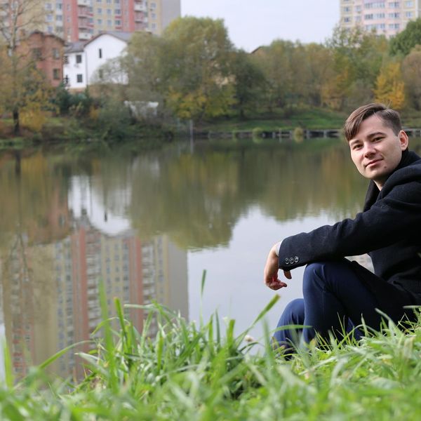 Smiling person sitting in a lotus pose outdoors near a lake.